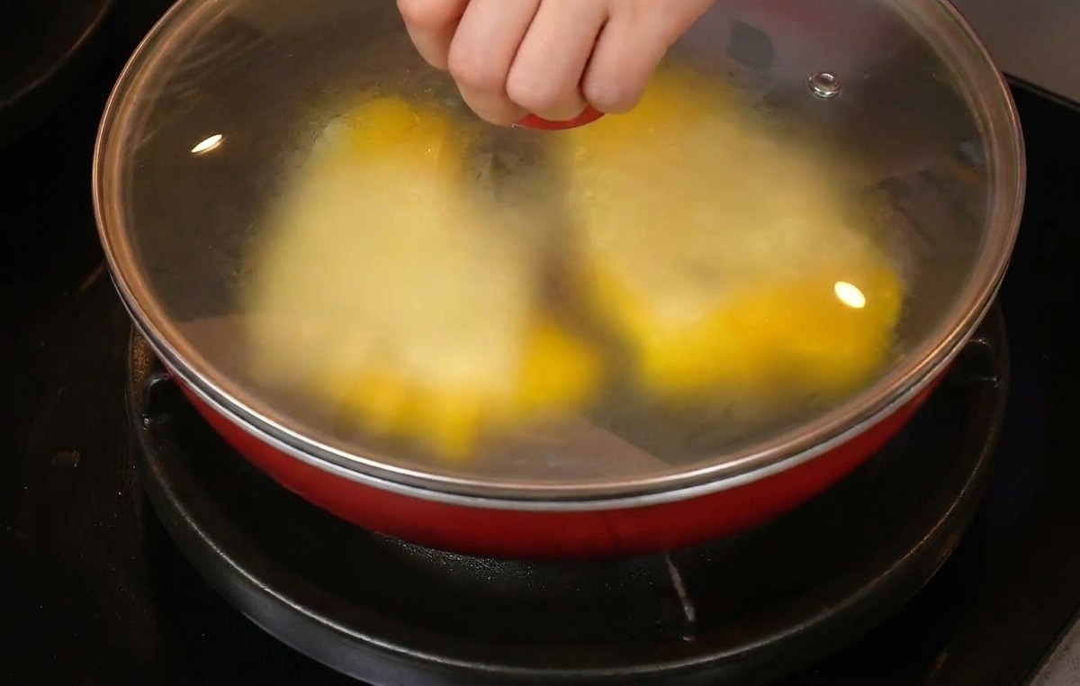 Una mano che posiziona un coperchio di vetro su una padella rossa con uova e formaggio.