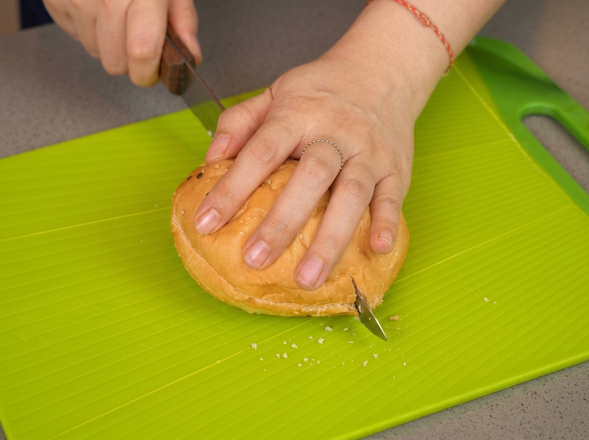 Mano che preme su un panino al sesamo mentre un coltello lo taglia a metà su un tagliere verde acido.