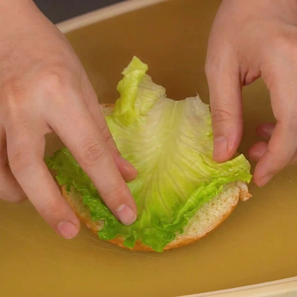 Mani che posizionano una foglia di lattuga verde brillante sulla base del panino.