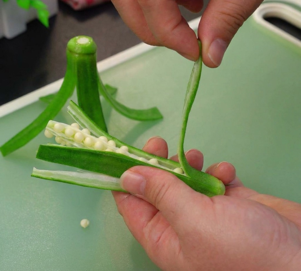 Mani che separano delicatamente le strisce esterne di un baccello di okra per rivelare e rimuovere il cuore bianco con i semi.