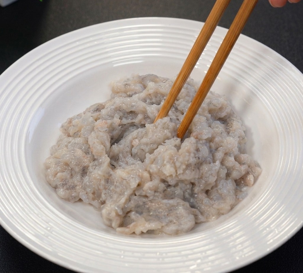 Miscelazione della pasta di gamberi crudi grigiastra in una ciotola di ceramica bianca con bacchette di legno.