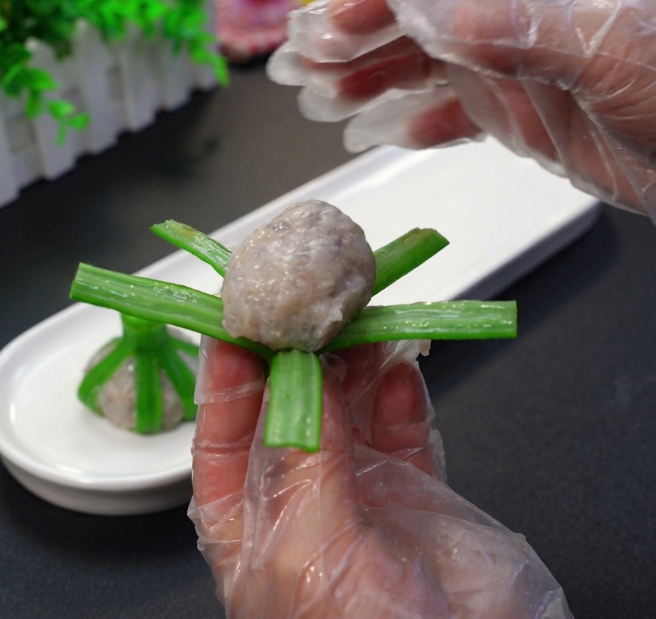 Mani guantate che modellano la pasta di gamberi crudi in una sfera e la avvolgono nelle strisce verdi di unokra sbollentata.