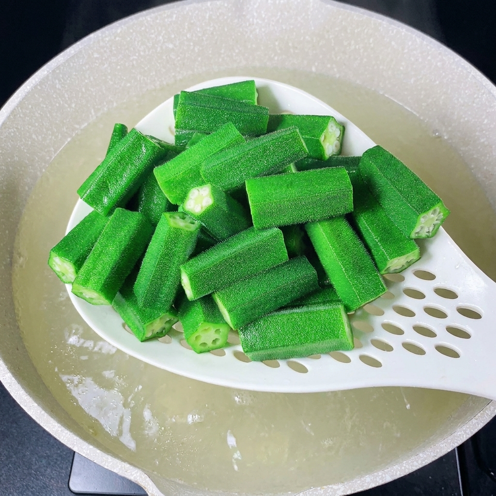 Una schiumarola bianca che solleva pezzi di okra appena sbollentati dallacqua bollente.
