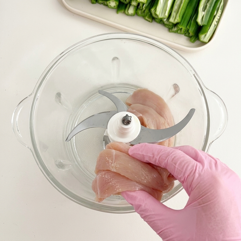 Mano con guanto che inserisce pezzi di pollo crudo in un robot da cucina in vetro con lama di metallo.