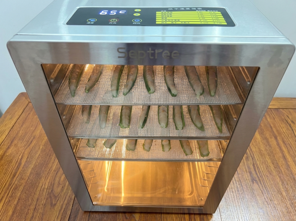 Vassoi carichi di okra ripiena inseriti in un essiccatore per alimenti in acciaio inossidabile illuminato.