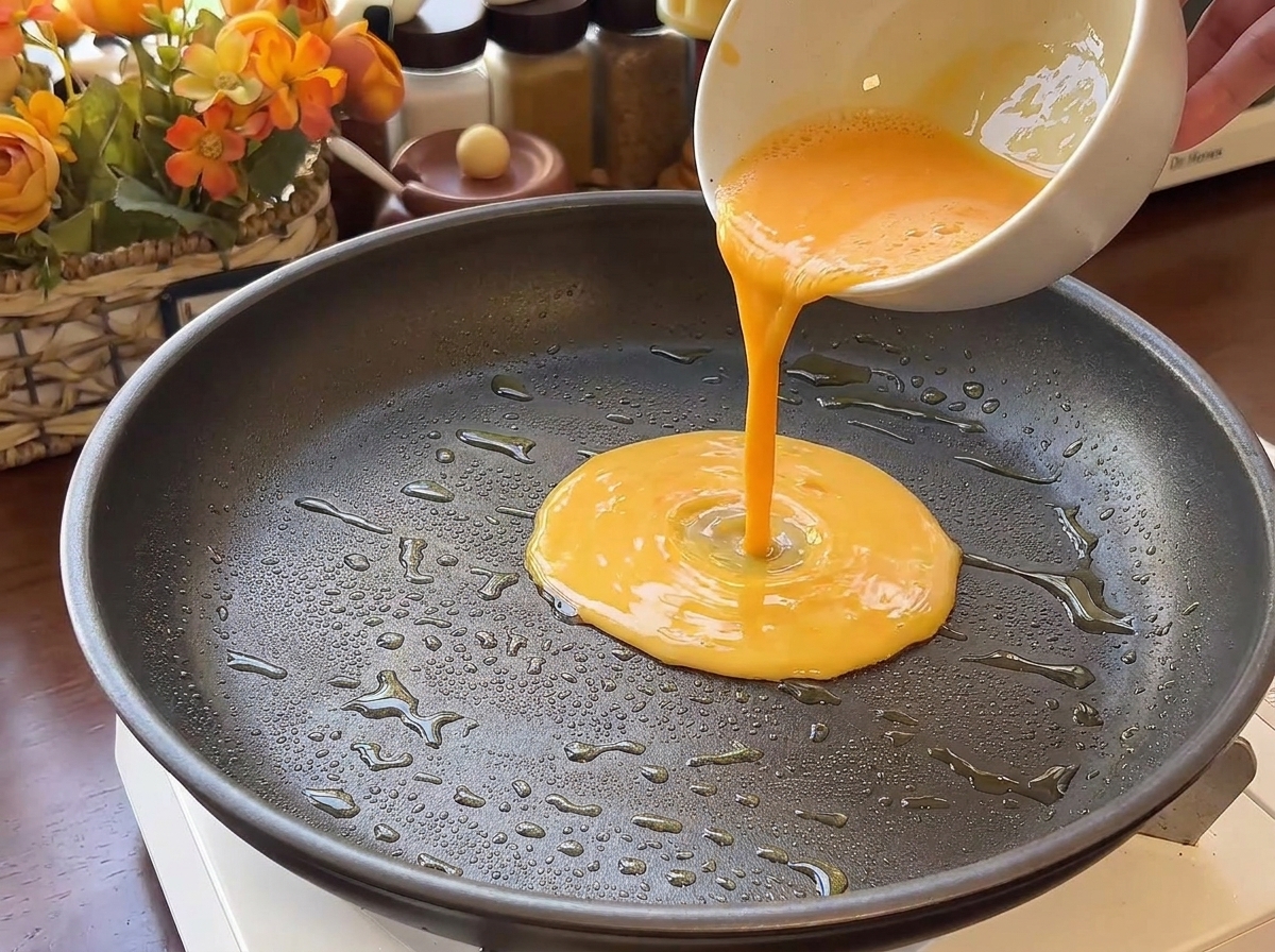 Liquido giallo delle uova versato in una padella antiaderente scura con gocce dolio visibili.