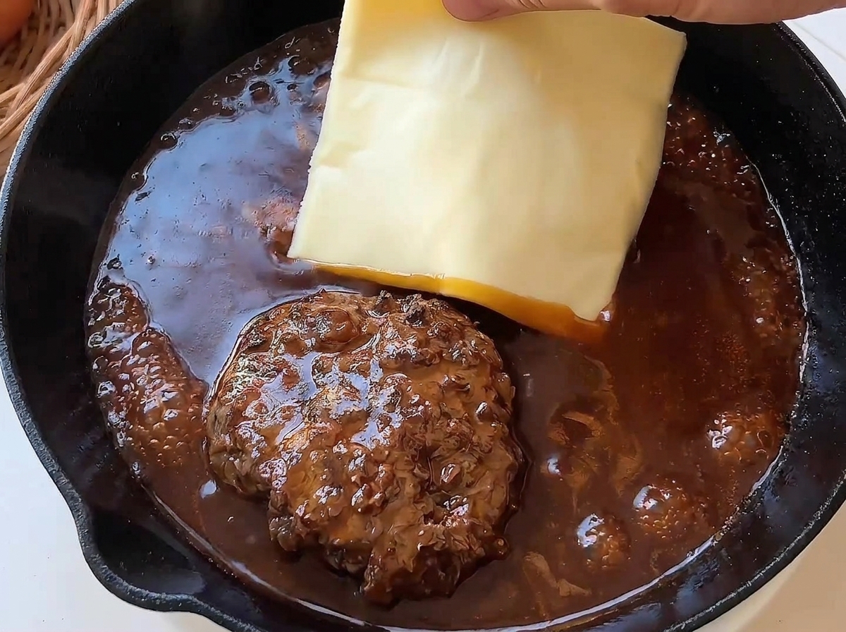 Una mano che appoggia una fetta di formaggio giallo sopra un hamburger che cuoce nella salsa bruna.