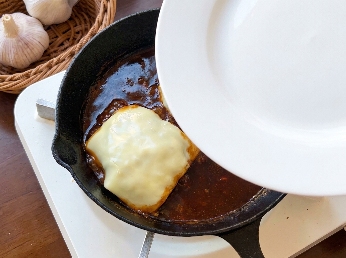 Un piatto bianco usato come coperchio sopra una padella di ghisa per far fondere il formaggio sulla carne.