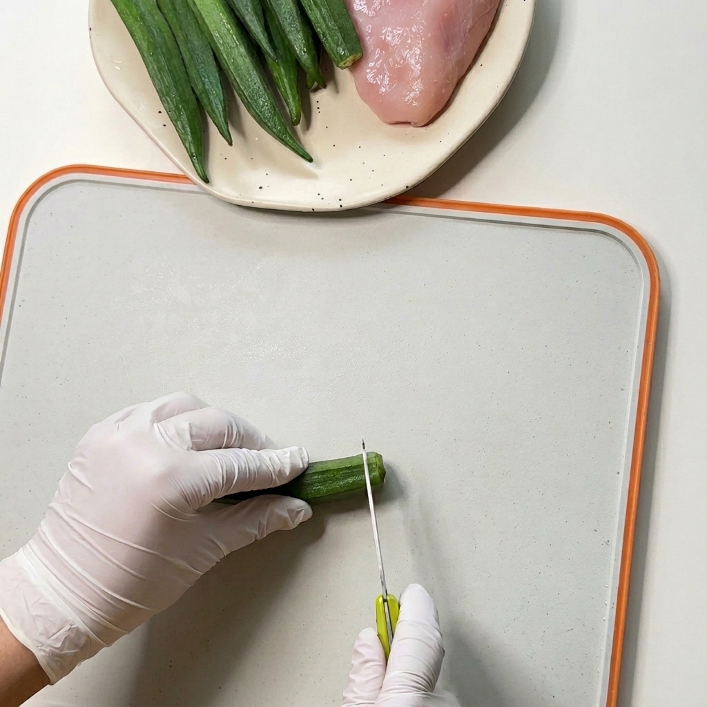 Mani con guanti usano un coltellino per tagliare le estremità di baccelli di okra verde su un tagliere bianco.