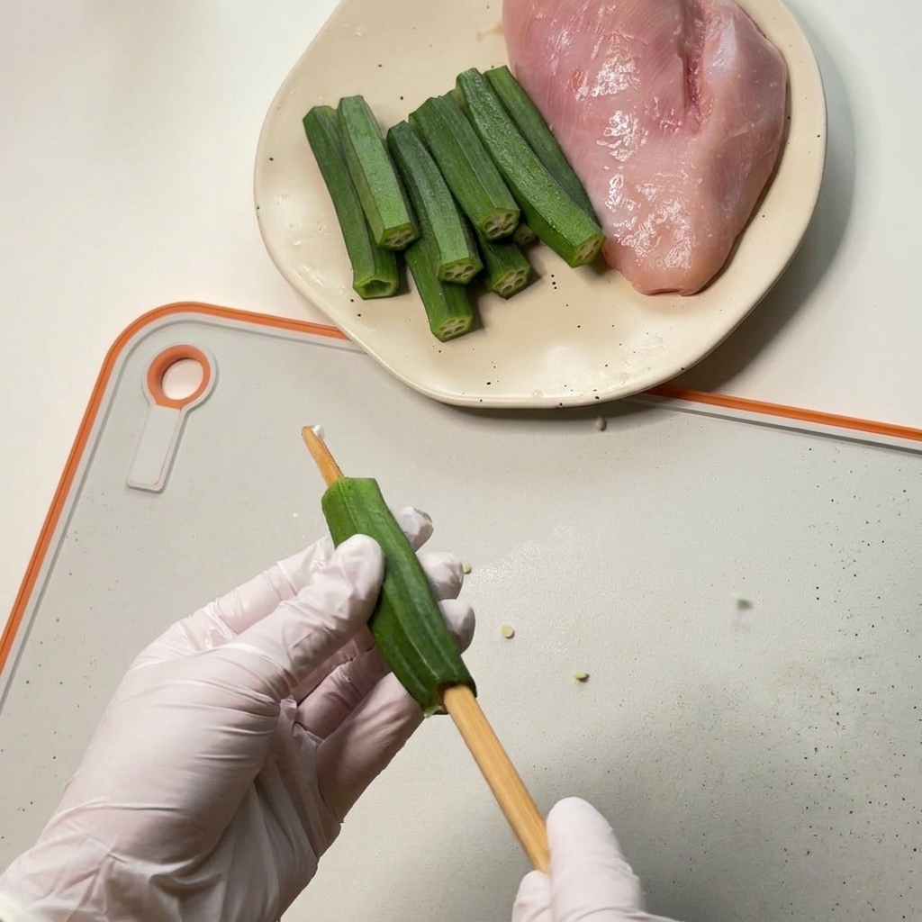 Mani con guanti svuotano il centro di un baccello di okra usando una bacchetta di legno spessa su un tagliere bianco.