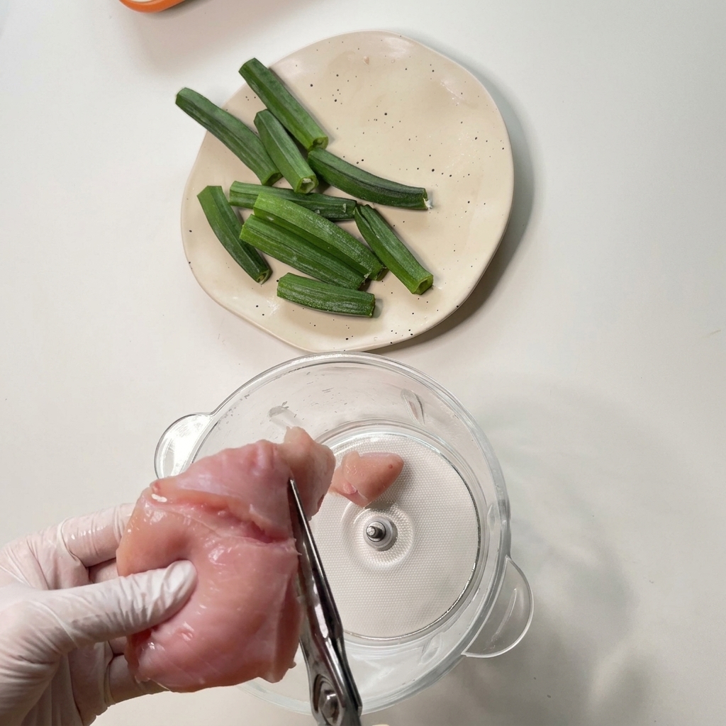 Mani con guanti usano forbici da cucina per tagliare un pezzo di petto di pollo crudo sopra la ciotola di vetro di un robot da cucina.