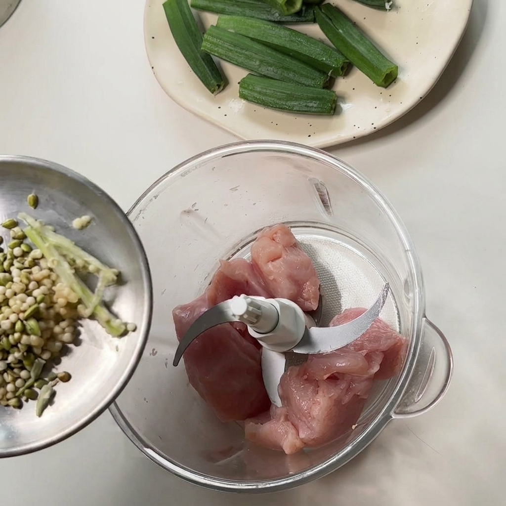 Versare semi e scarti di okra da un piattino di metallo in un robot da cucina contenente pezzi di pollo crudo.