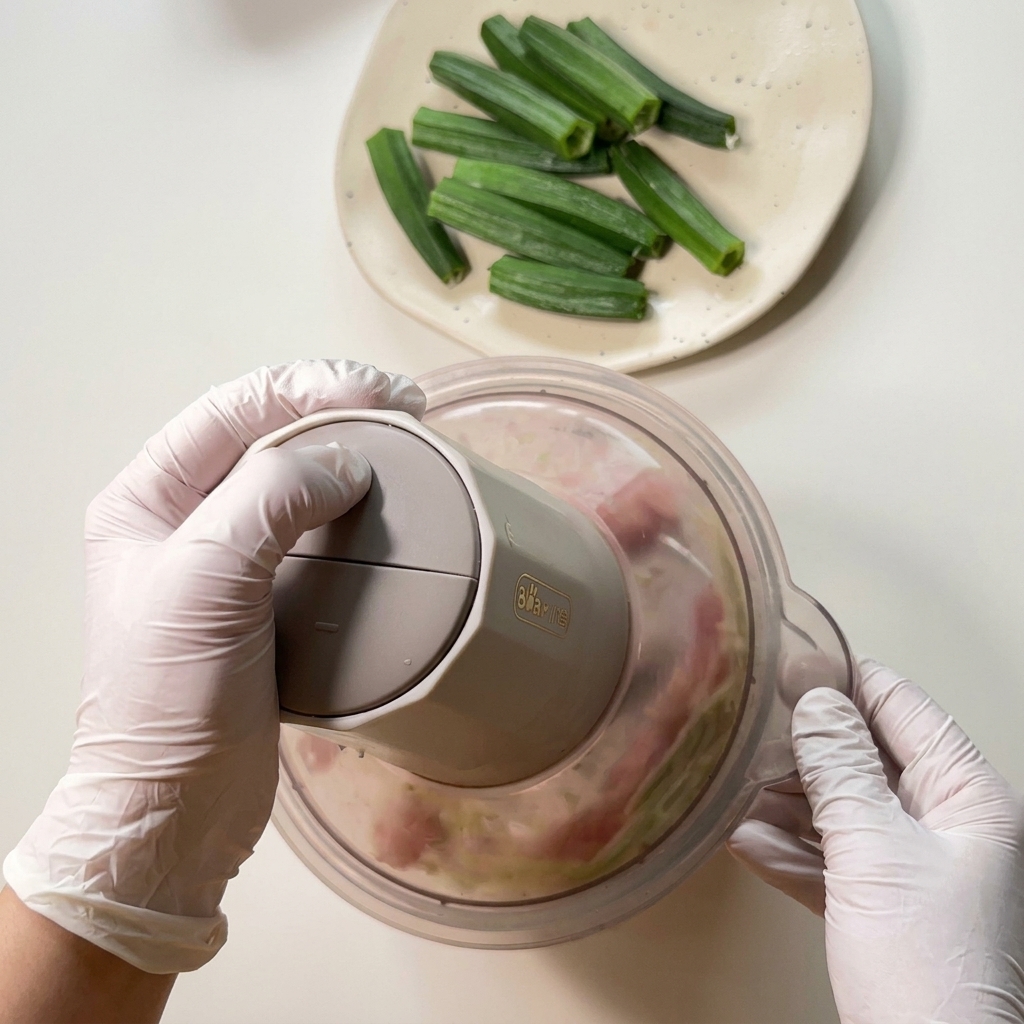 Mani con guanti premono su un mini robot da cucina per frullare pollo crudo e scarti di okra verde fino a ottenere una pasta.