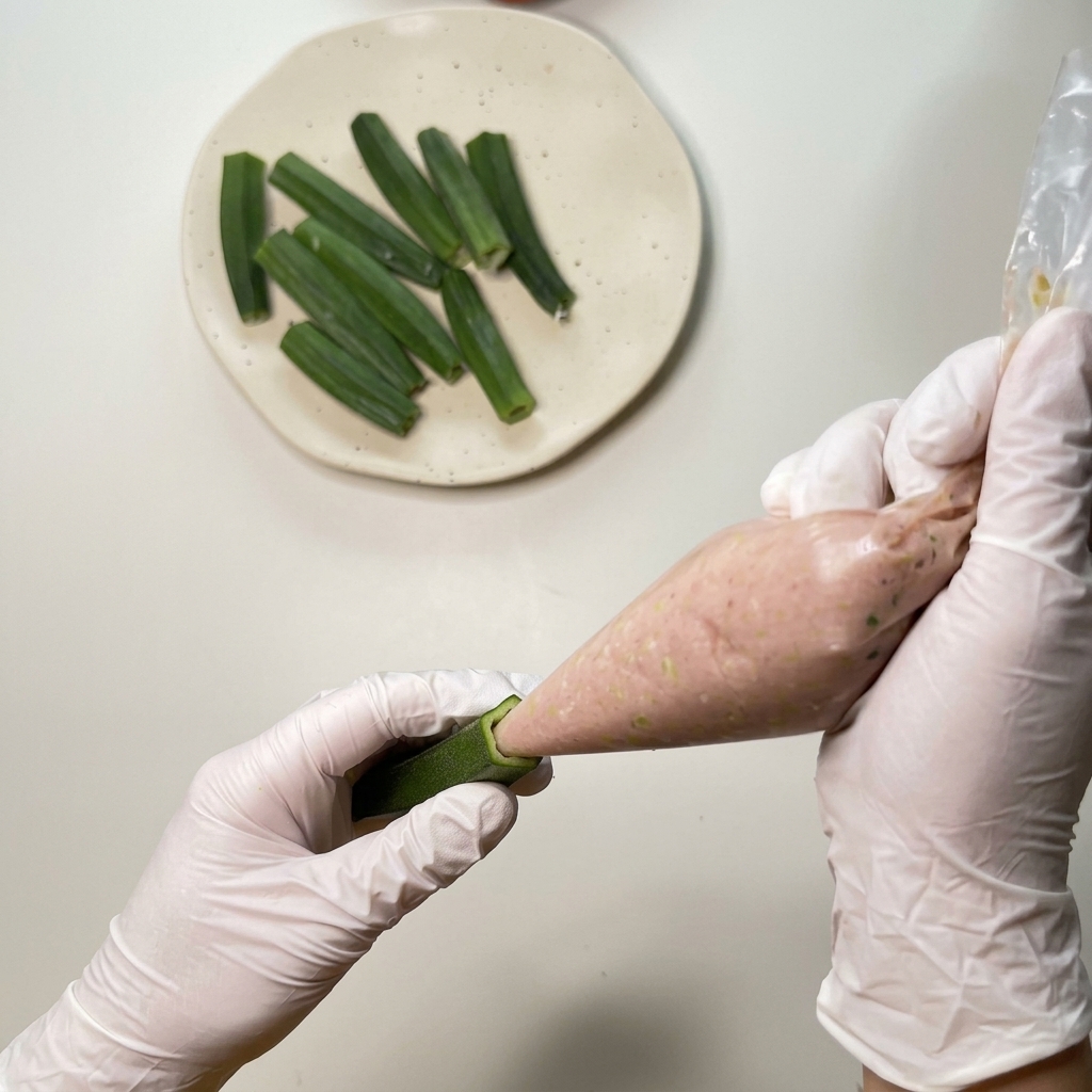 Mani con guanti usano una sac à poche di plastica per inserire il ripieno di pollo tritato in un baccello di okra svuotato.
