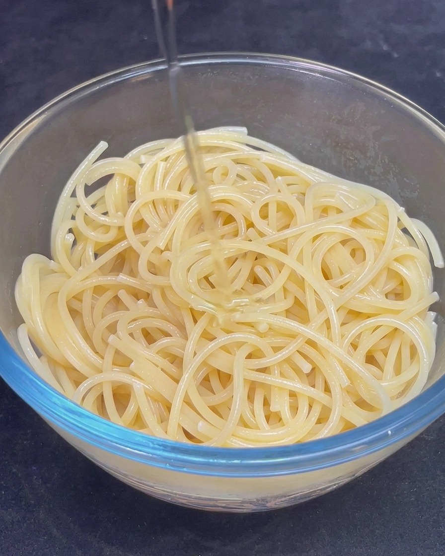 Un filo dolio doliva dorato versato sopra una ciotola di vetro piena di spaghetti cotti.