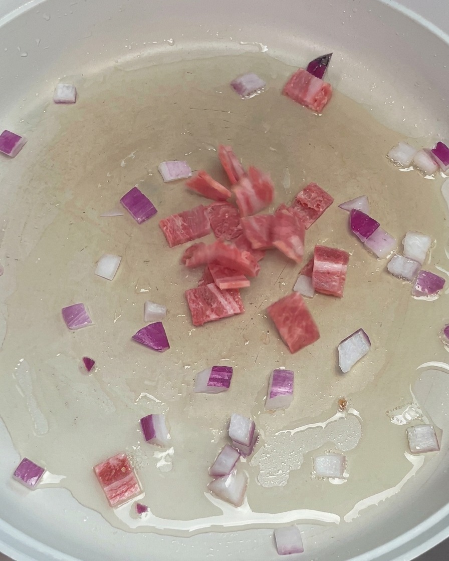 Bacon a cubetti e cipolla rossa tritata che sfrigolano in olio caldo in una padella bianca.