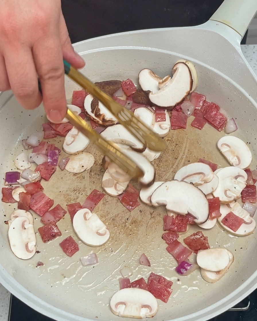 Una mano che usa le pinze per mescolare funghi, cipolla e bacon in una padella bianca.