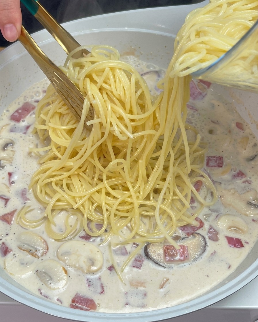 Pinze che adagiano gli spaghetti cotti nella salsa cremosa bollente con funghi e bacon.