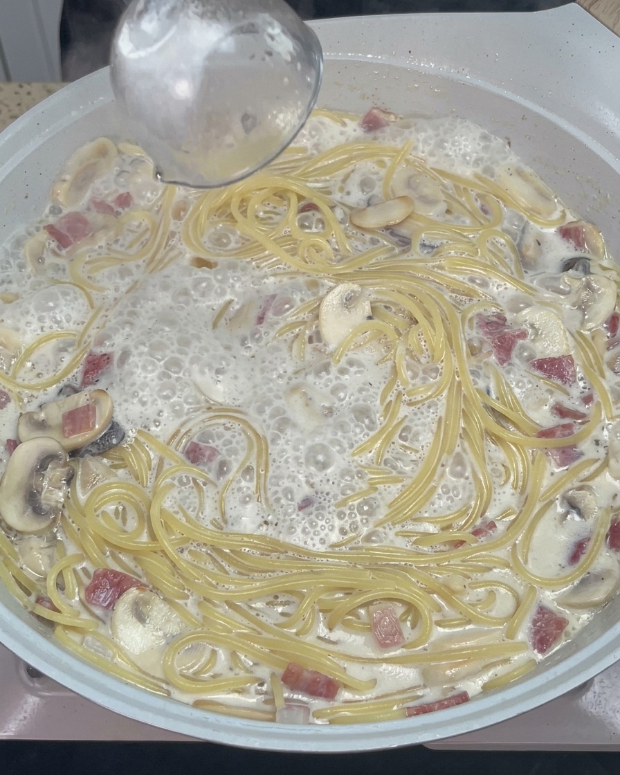 Cucchiaio di legno che mescola spaghetti cremosi con formaggio fuso e condimenti in padella.