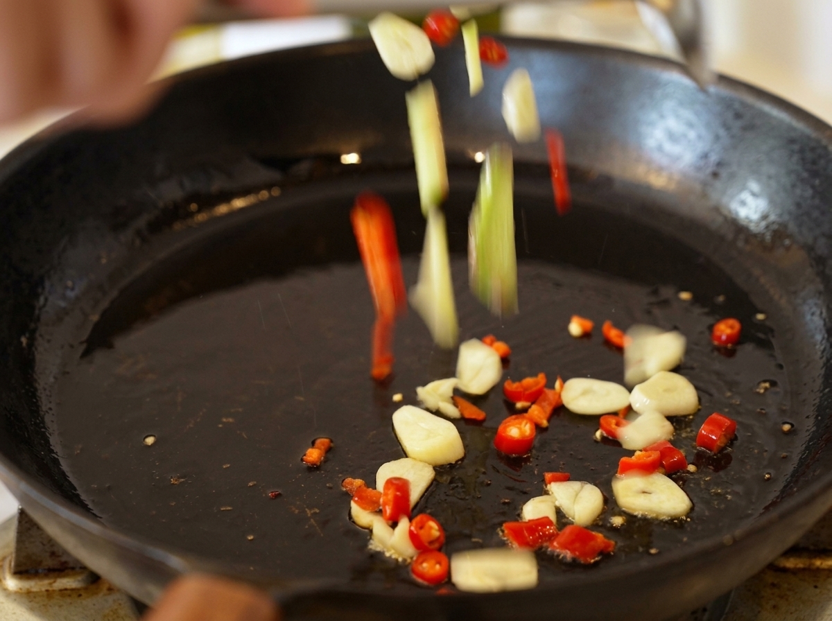 Aglio affettato e pezzi di peperoncino che cadono in una padella nera con olio doliva caldo.