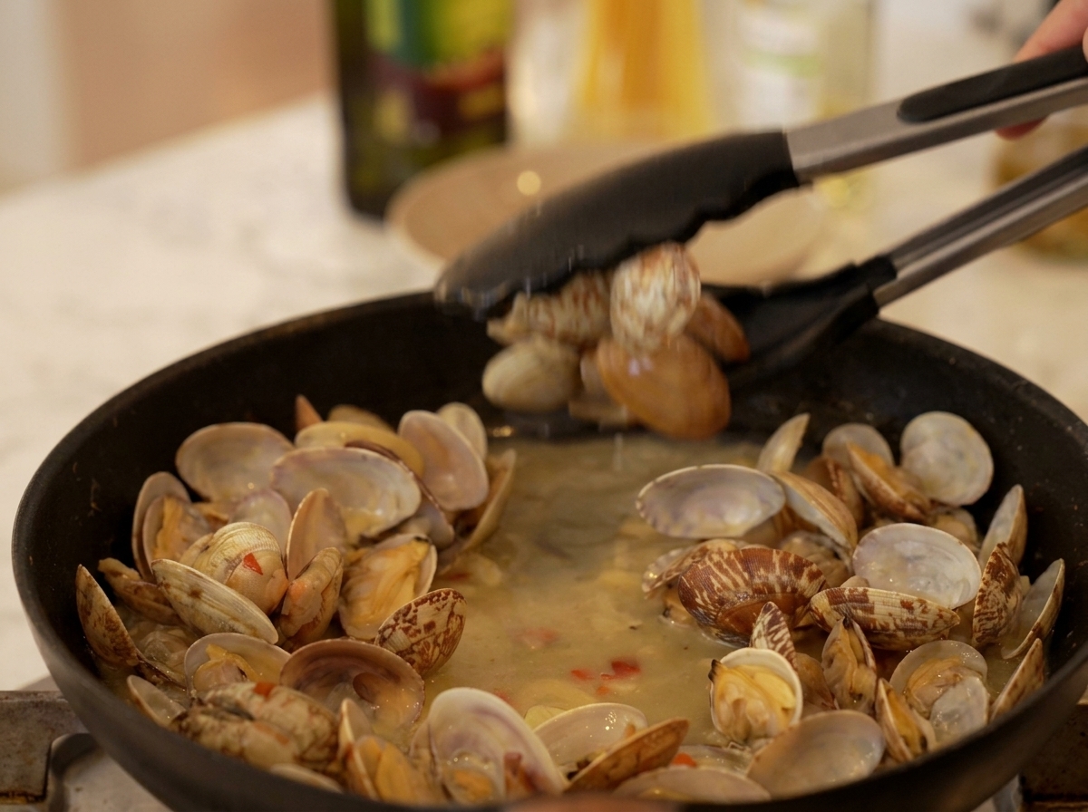Pinze da cucina che prelevano le vongole già aperte da un sugo bollente in una padella nera.