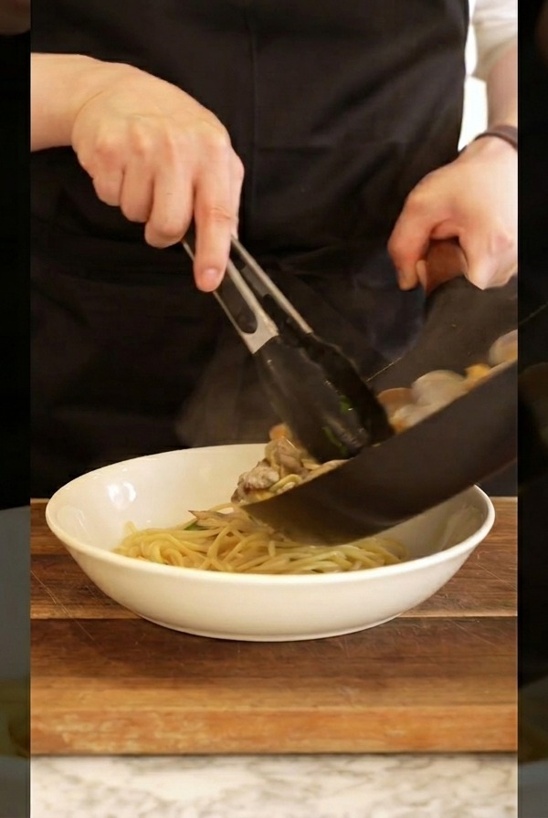 Un cuoco che usa le pinze per trasferire gli spaghetti alle vongole caldi dalla padella a una ciotola bianca.