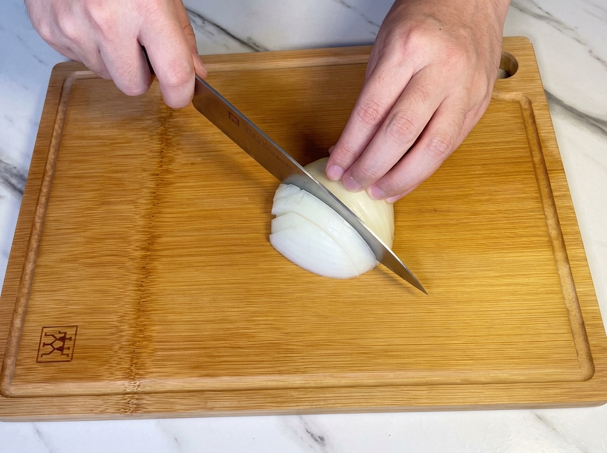 Mani che usano un coltello da chef per tritare mezza cipolla bianca su un tagliere di legno.