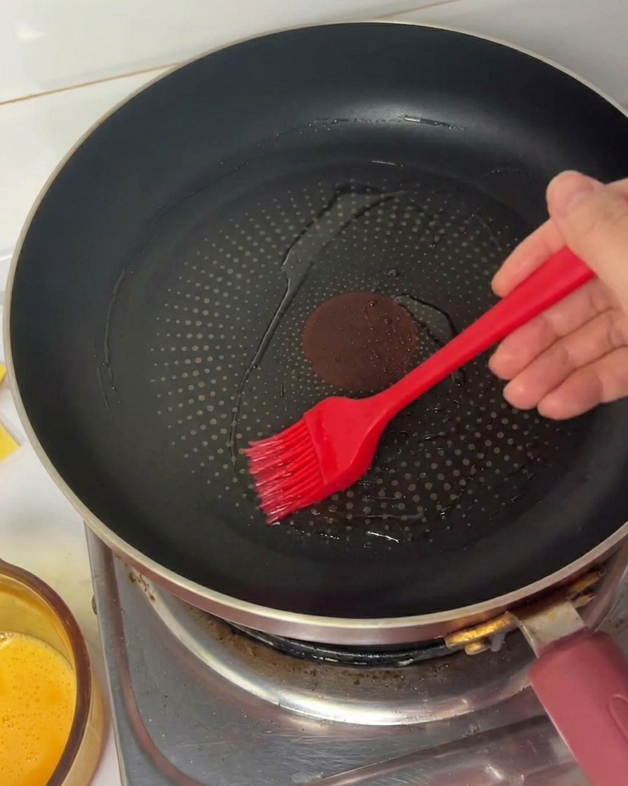 Una mano che usa un pennello in silicone rosso per spargere olio in una padella antiaderente nera.