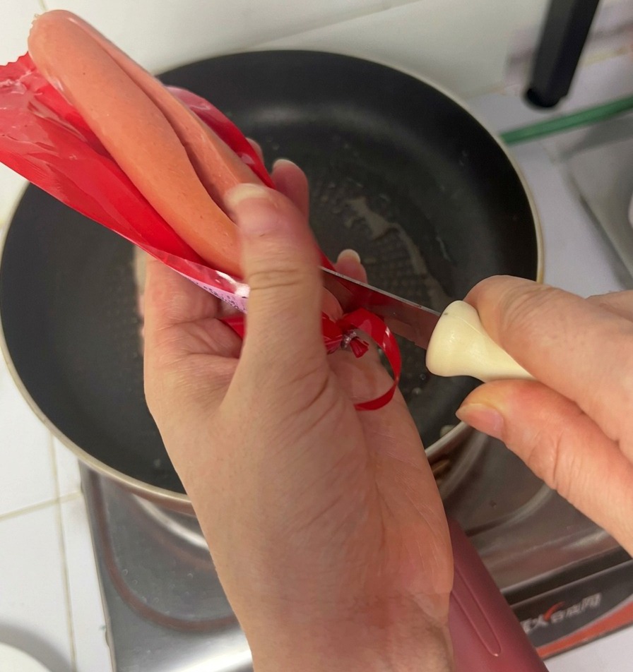 Mani che aprono una confezione di würstel di prosciutto sopra una padella con bacon in cottura.