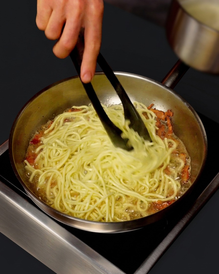 Un pentolino che versa acqua calda della pasta in una padella con spaghetti e guanciale mentre vengono saltati con le pinze.