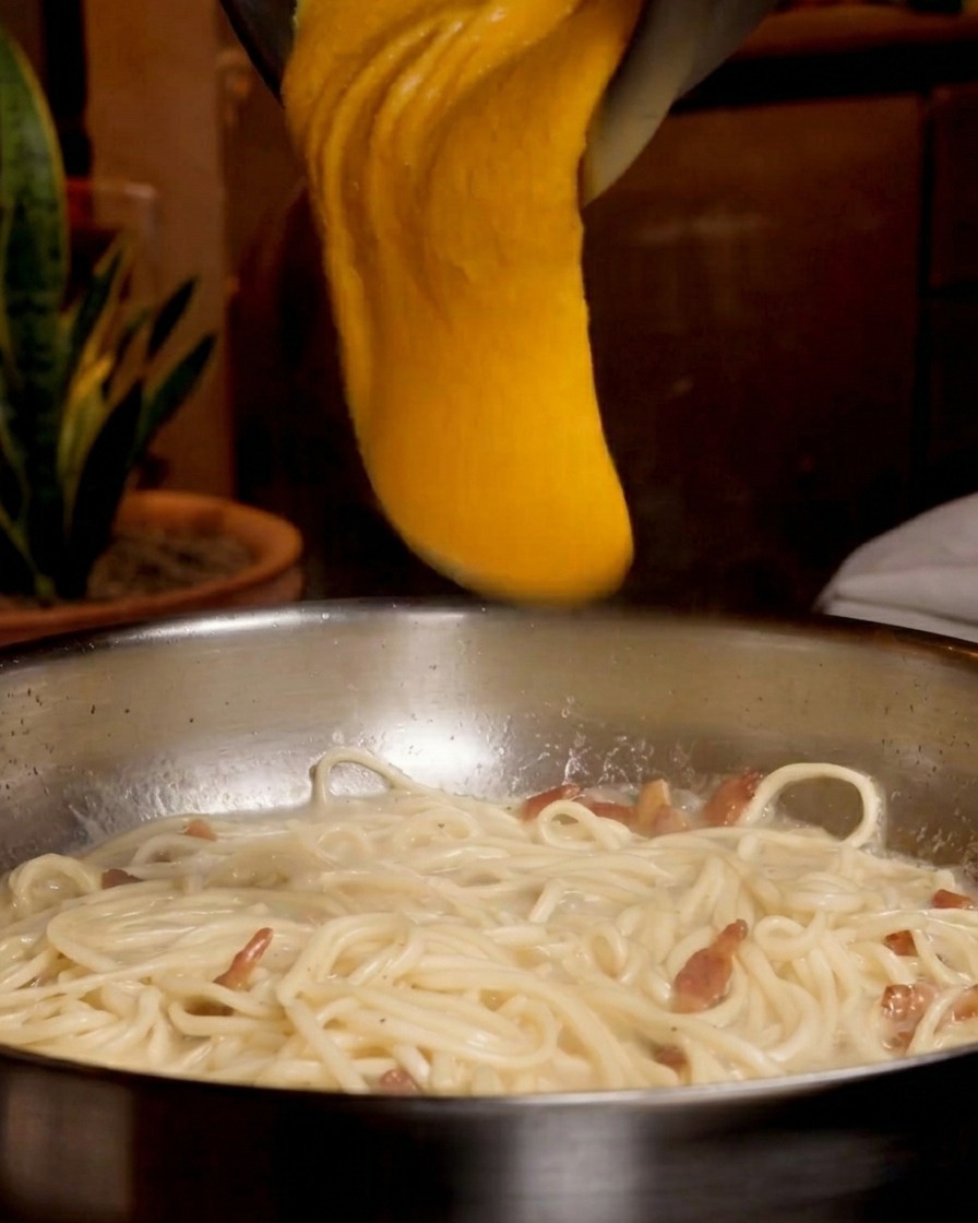 Un denso composto arancione brillante di uova e formaggio versato da una ciotola di metallo sopra una padella con spaghetti e guanciale.