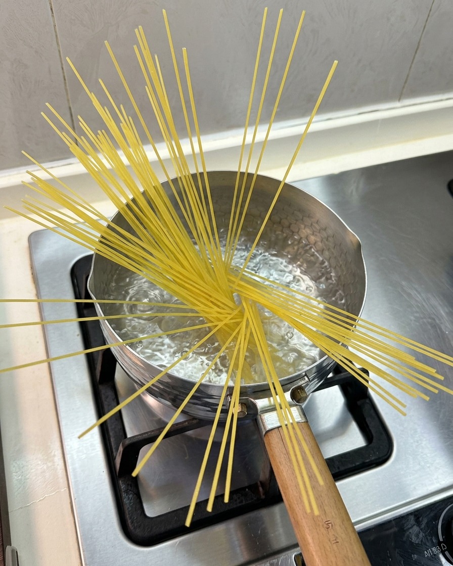 Spaghetti secchi aperti a ventaglio e parzialmente immersi in una piccola pentola di acqua bollente su un fornello a gas.