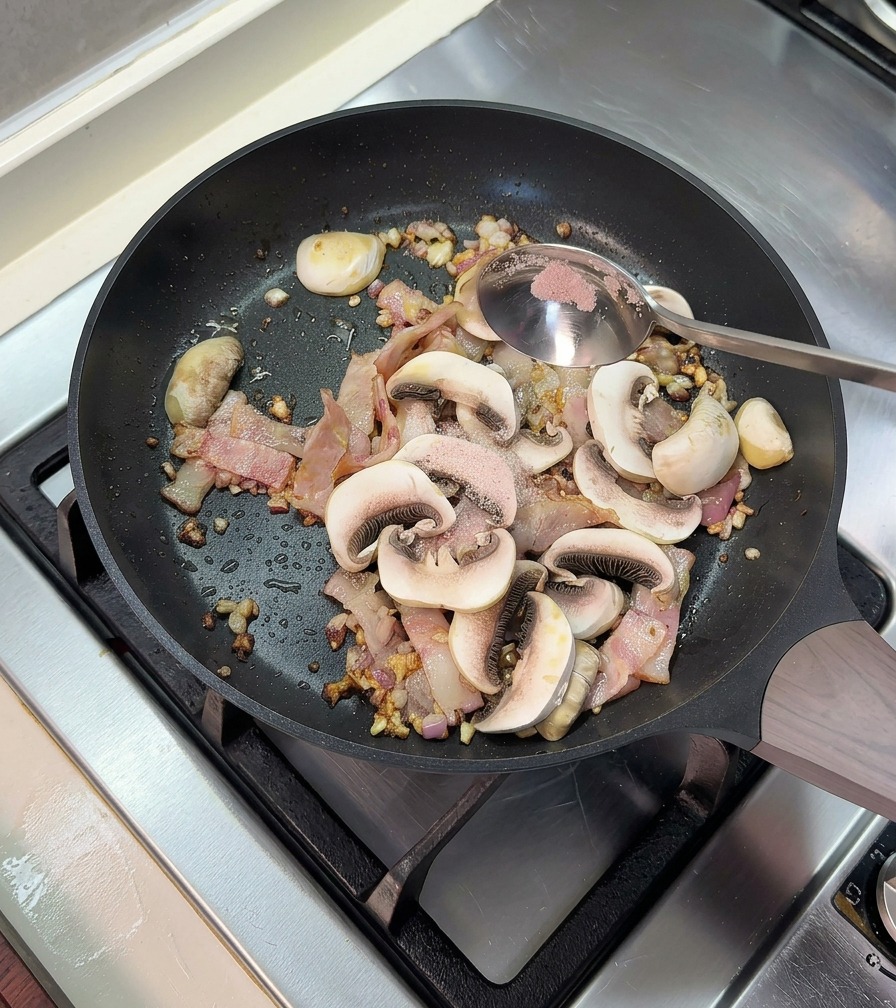 Aggiunta di un cucchiaio di sale rosa in una padella con pancetta cotta e funghi affettati.