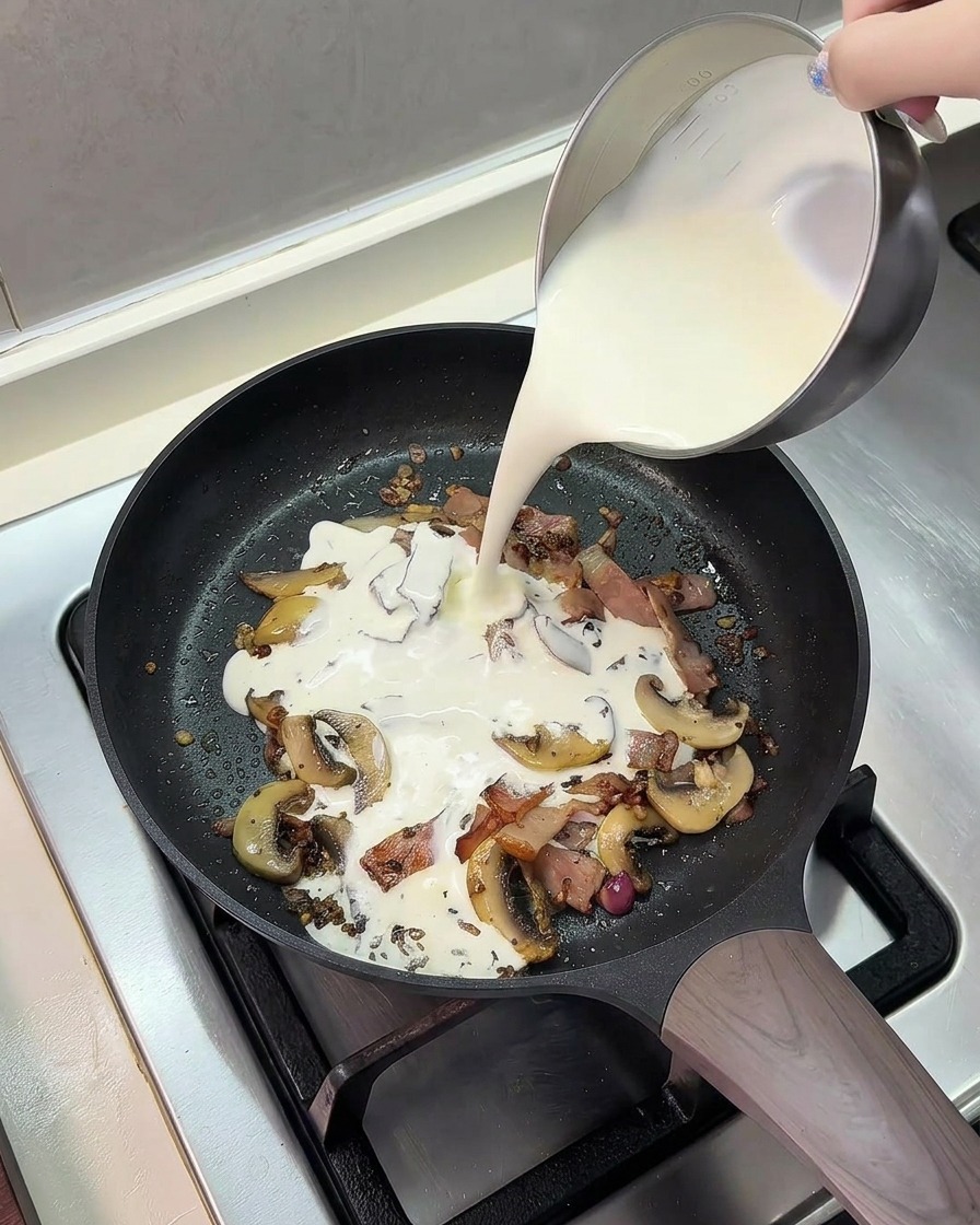 Versare una miscela di latte e panna fresca da una lattiera di metallo in una padella con pancetta e funghi.