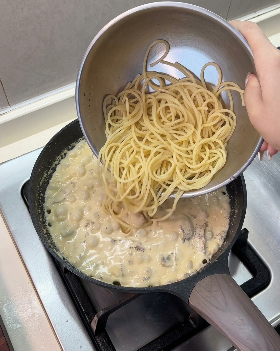 Spaghetti cotti versati da una ciotola in acciaio inossidabile in una padella con salsa cremosa che sobbolle.