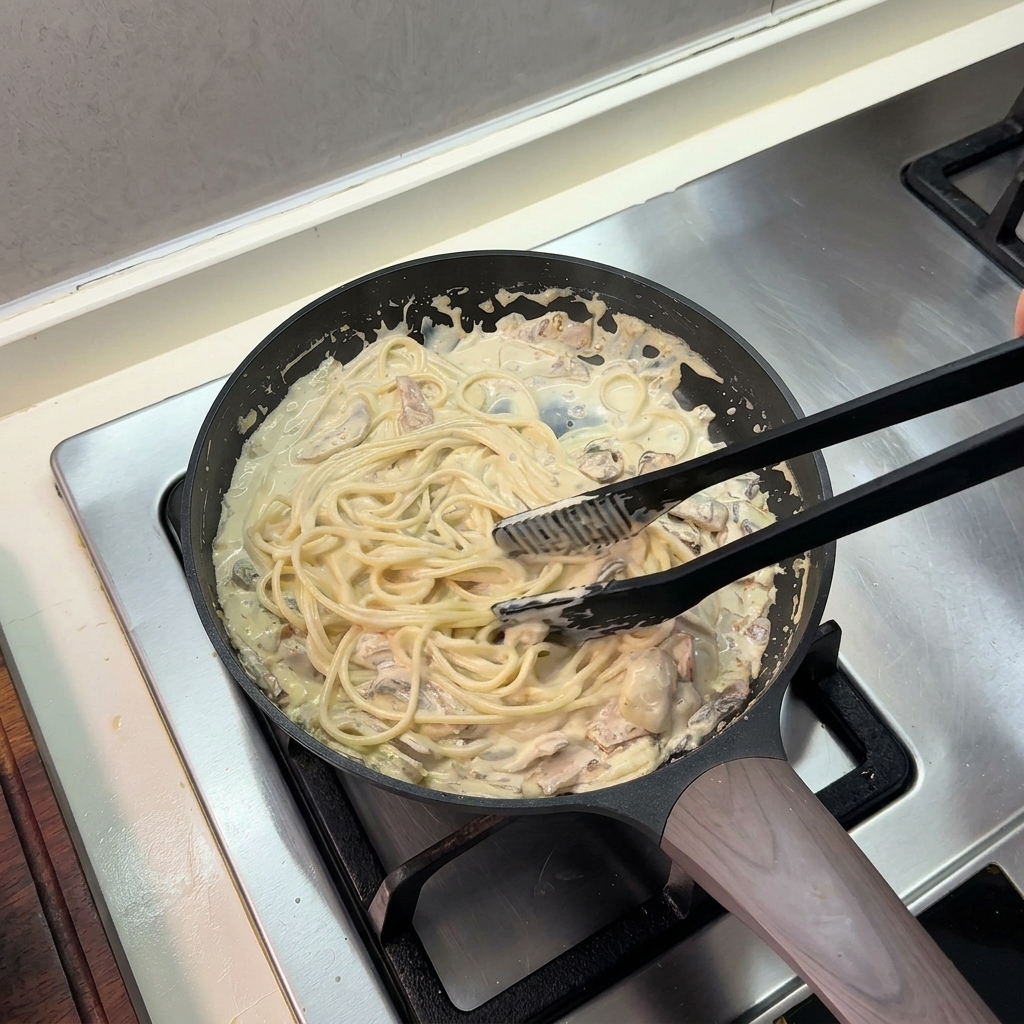 Pinze da cucina nere che saltano gli spaghetti in una ricca salsa cremosa allinterno di una padella.