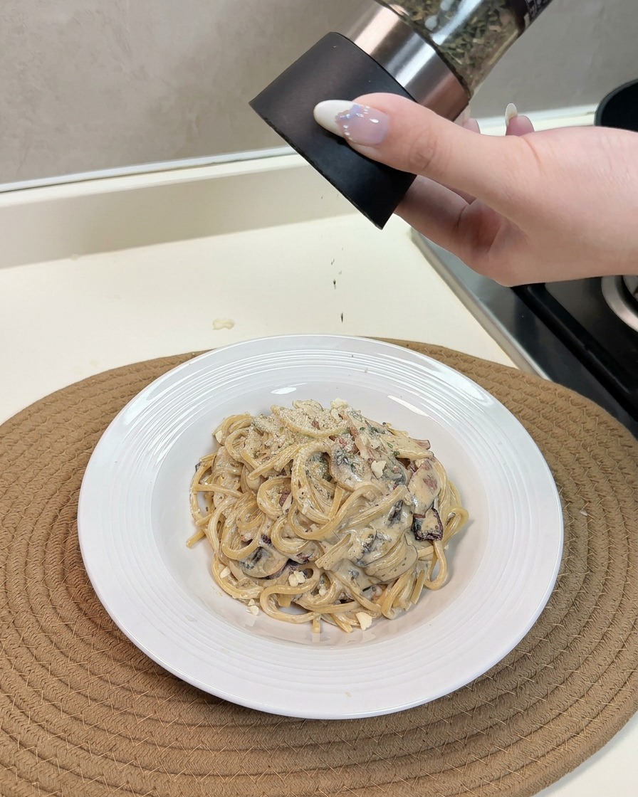 Una mano che utilizza una grattugia per spolverare formaggio ed erbe aromatiche su un piatto caldo di spaghetti cremosi ai funghi e pancetta.