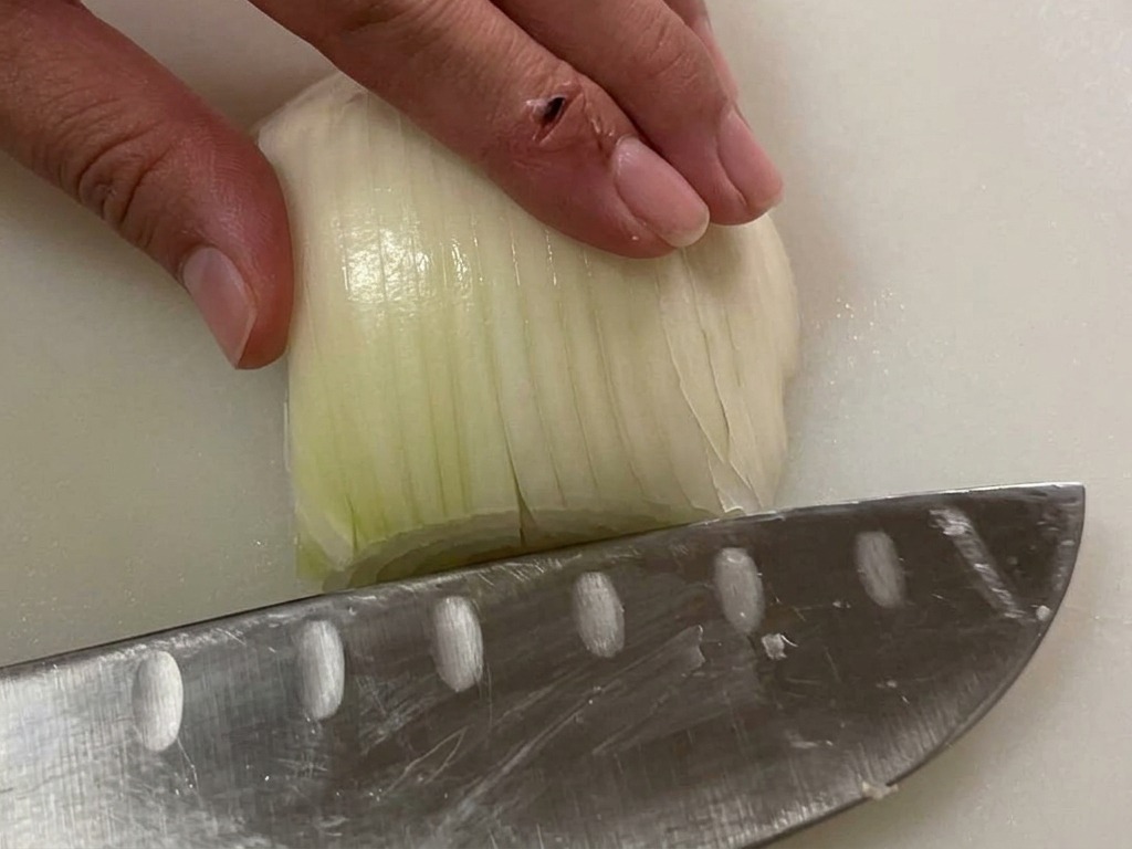 Mano che tiene mezza cipolla bianca su un tagliere mentre un coltello prepara il trito.
