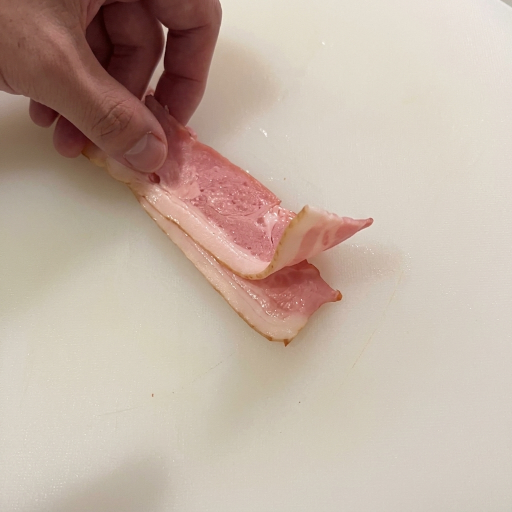 Mano che ripiega due strisce di pancetta cruda su un tagliere, pronta per essere tagliata.