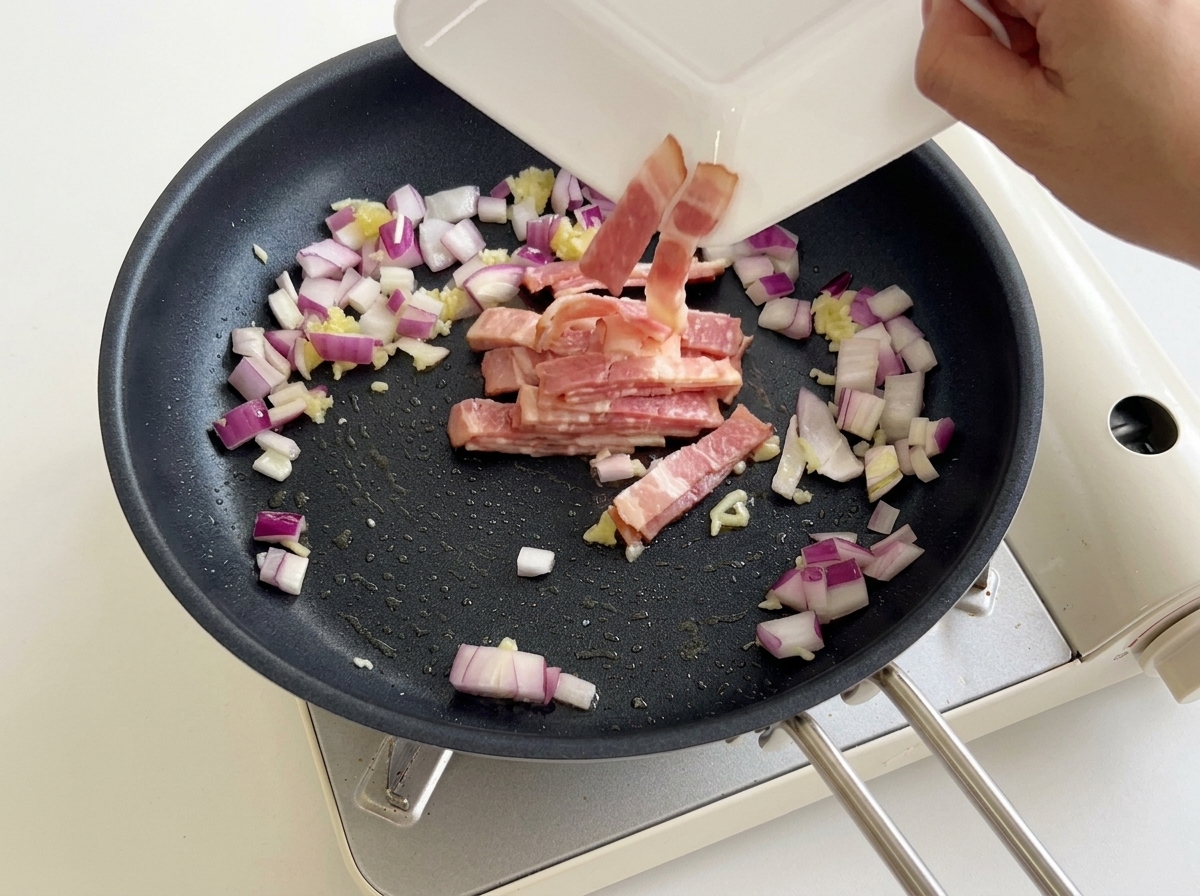Pancetta tagliata versata da un piatto bianco in padella con cipolla e aglio.