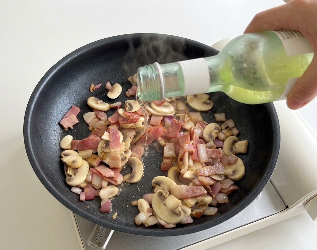 Mano che versa vino bianco da una bottiglietta verde in una padella calda con funghi e pancetta.