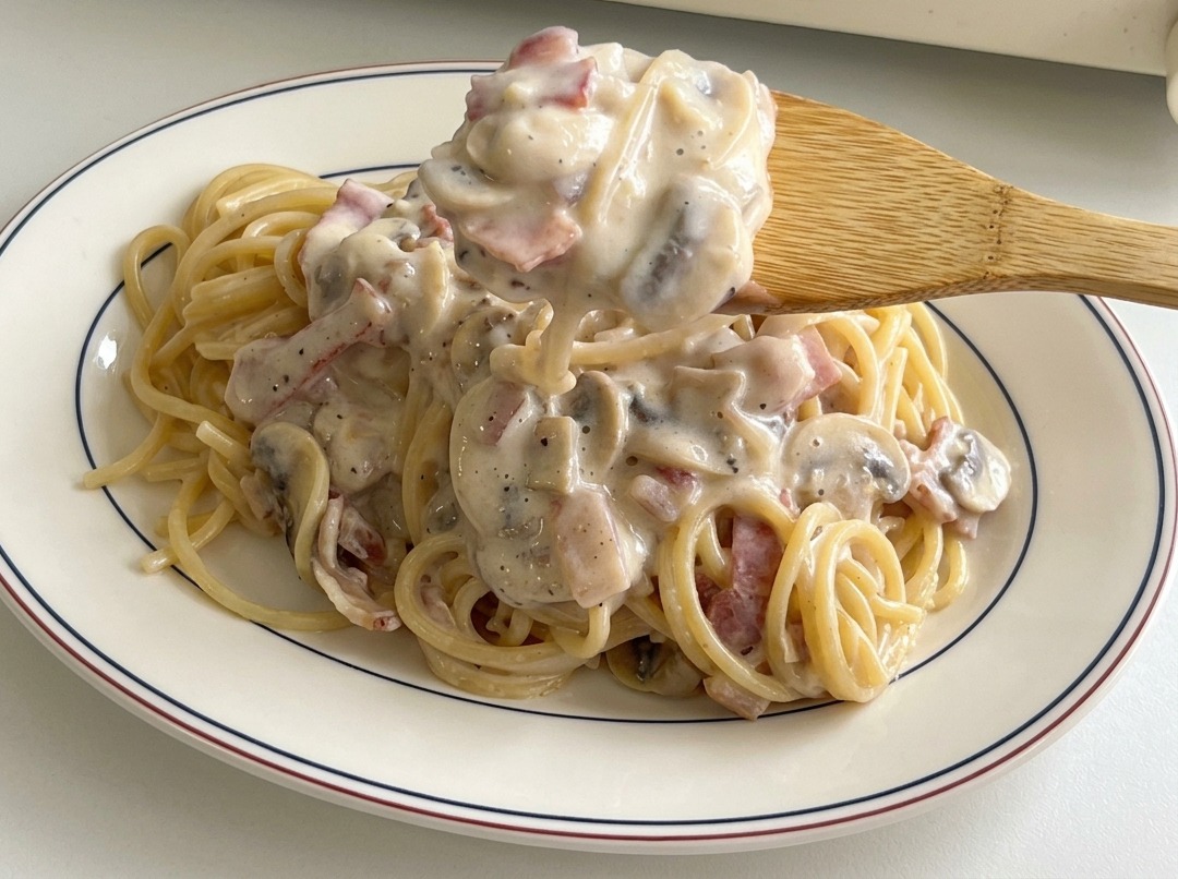Cucchiaio di legno che versa salsa cremosa con funghi e pancetta su una porzione di spaghetti.