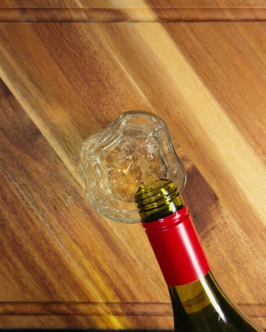 Vino bianco versato da una bottiglia di vetro verde in un bicchiere trasparente su una superficie di legno.