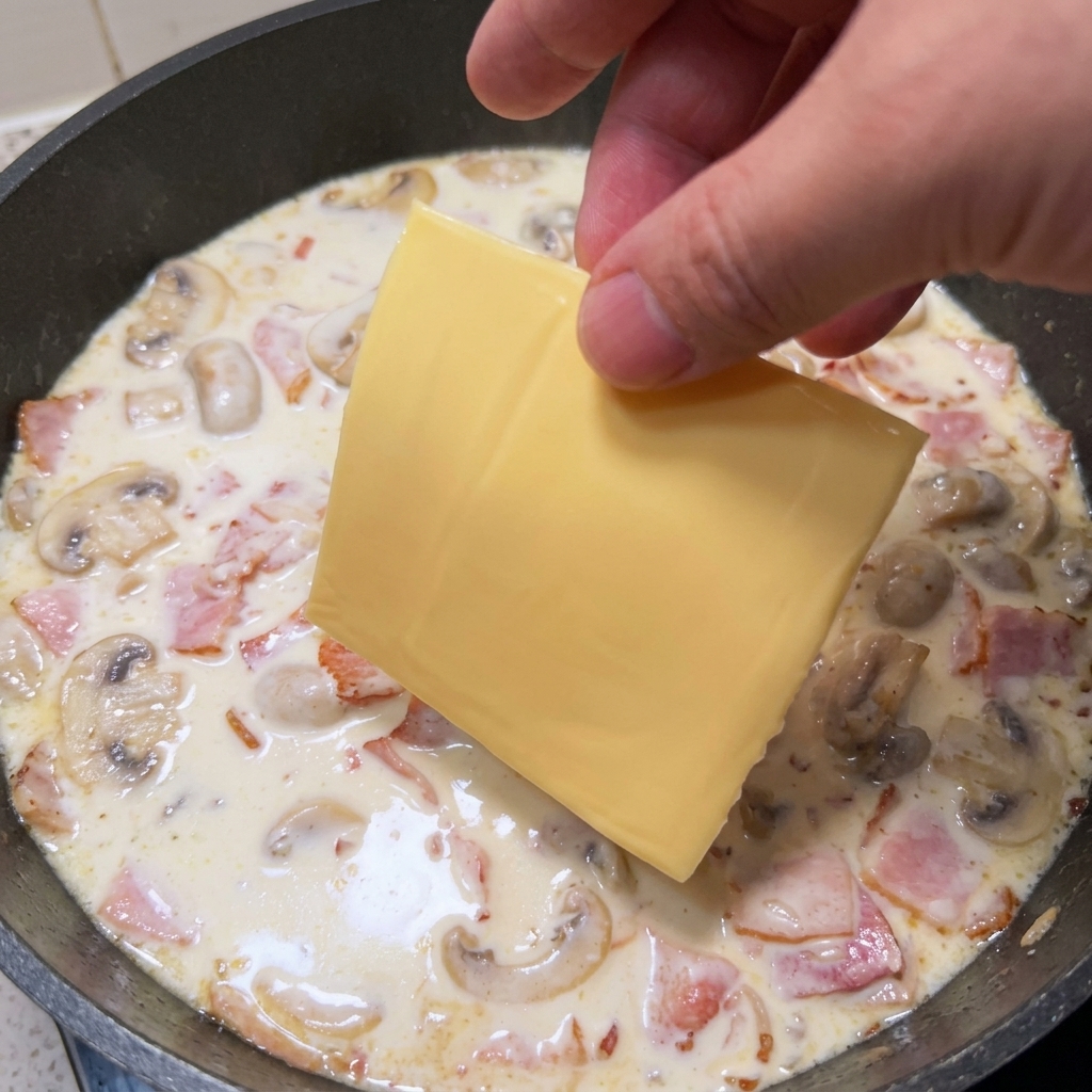 Una mano posiziona una fetta di formaggio giallo nella salsa cremosa in padella.