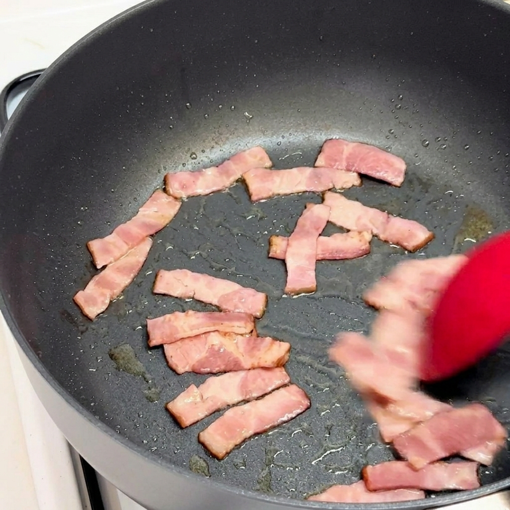 Pezzi di pancetta saltati in padella con una spatola di silicone rossa.