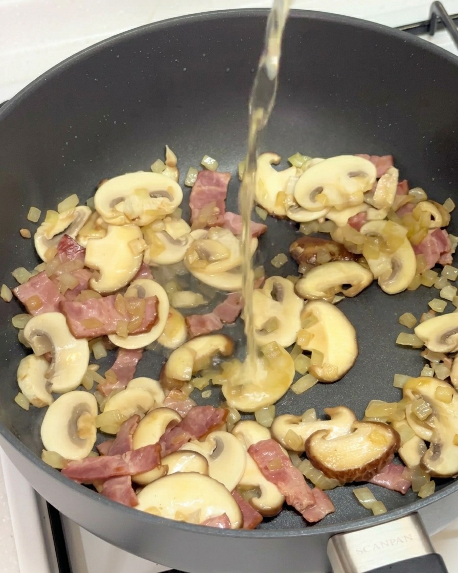 Liquido trasparente versato in una padella con funghi, pancetta e cipolla saltati.