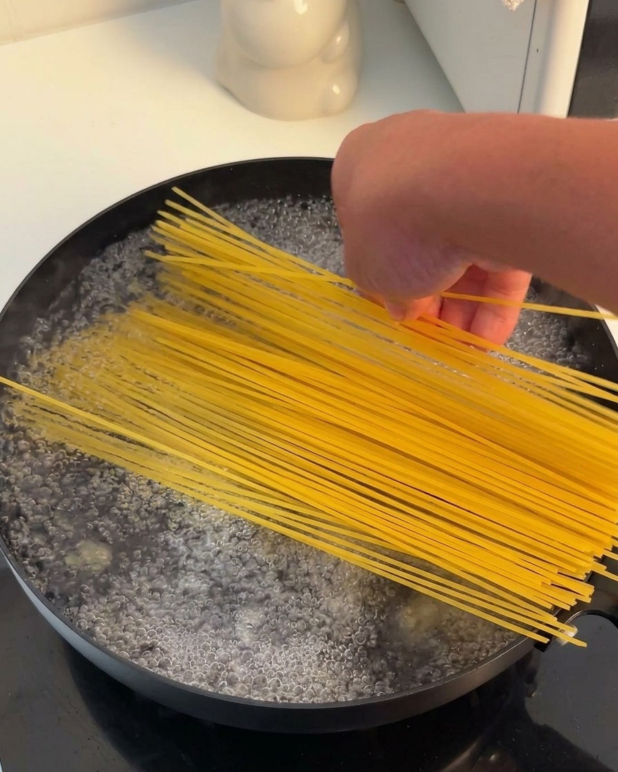 Una mano che mette un fascio di spaghetti gialli secchi in una pentola nera piena di acqua bollente.