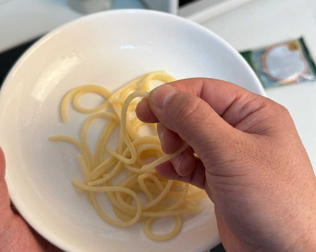 Una mano che pizzica uno spaghetto cotto sopra una ciotola bianca per controllarne la consistenza.