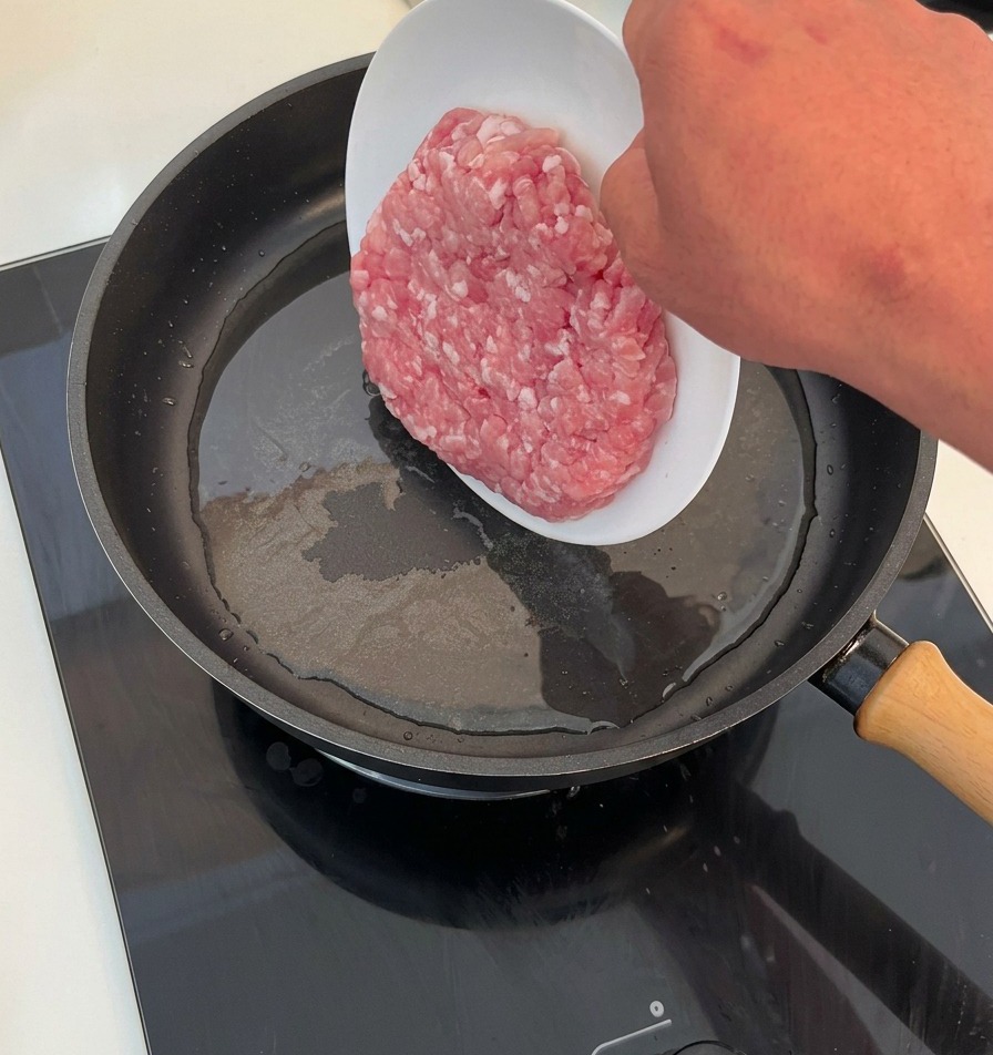 Una mano che fa scivolare una porzione di carne macinata cruda da un piatto bianco in una padella calda con olio.