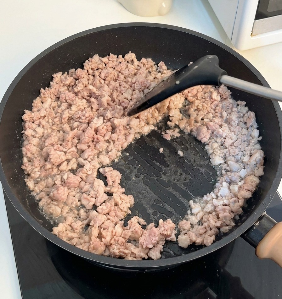 Una spatola nera che mescola e sgrana la carne macinata rosolata in una padella scura.
